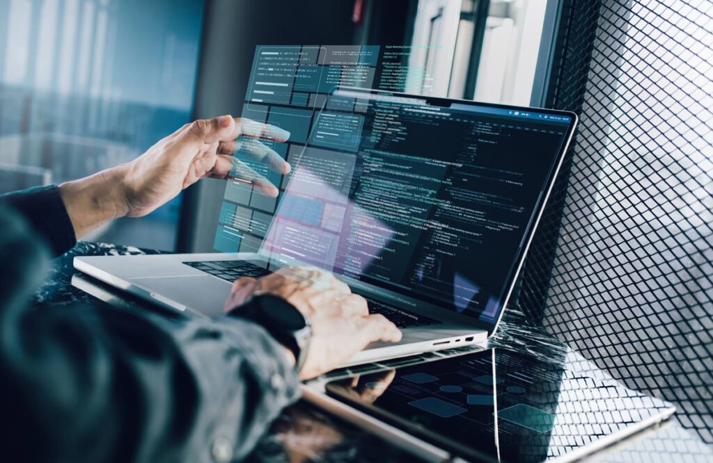 Side view of a person’s hands working on a laptop in a modern office. A holographic, semi-transparent window displaying lines of code and data modules is superimposed over the laptop screen and keyboard.
