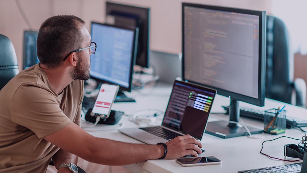 A man wearing over-ear headphones works at a modern workstation. He sits before a laptop and three large external monitors, all displaying complex lines of code. A smartphone is propped up on the desk, which also holds a mouse and multiple mousepads, creating a focused, high-tech programming environment.