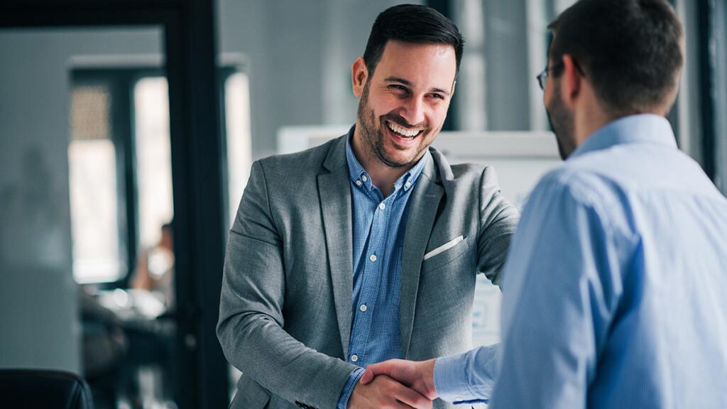A man in a grey blazer and blue button-down shirt smiles warmly while shaking hands with another person in a light blue shirt. The scene takes place in a bright, modern office environment with a shallow depth of field, focusing on the greeting and professional connection.