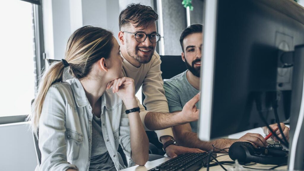 To round out your collection, here is the alt text for the final image: Alt Text: Three young professionals—two men and one woman—collaborate in a bright, modern office. The woman is seated, while the two men lean in behind her, all focusing on a computer monitor that is partially visible in the foreground. They appear engaged and are smiling during their discussion.