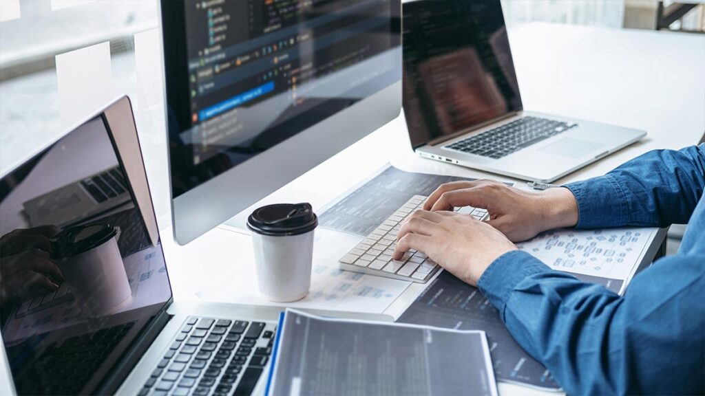 A software developer's hands typing on a wireless keyboard at a white desk. The workstation features a large desktop monitor and two laptops, all displaying lines of programming code and development interfaces, with a coffee cup nearby in a bright, modern office.