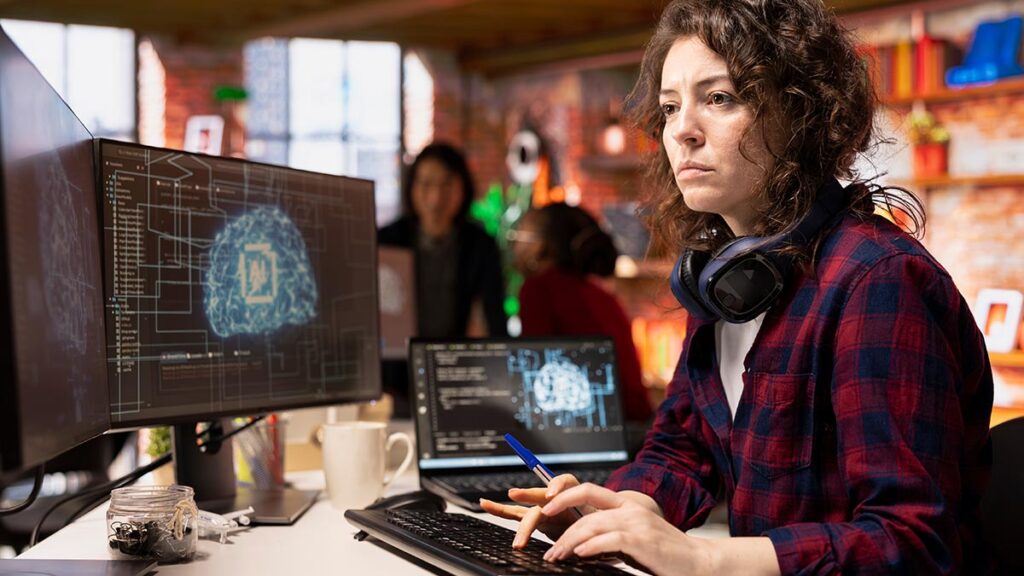 Focused female developer working in a high-tech office environment. She is typing on a keyboard while looking at a large monitor featuring a 3D digital brain model. The setting features warm, ambient lighting and industrial decor.