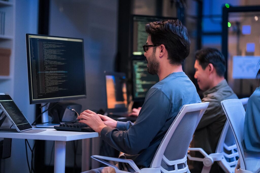 A software engineer wearing glasses works on a desktop computer displaying lines of code in a dimly lit, modern office. Other team members are visible in the background working at their respective stations, highlighting a collaborative tech development environment.