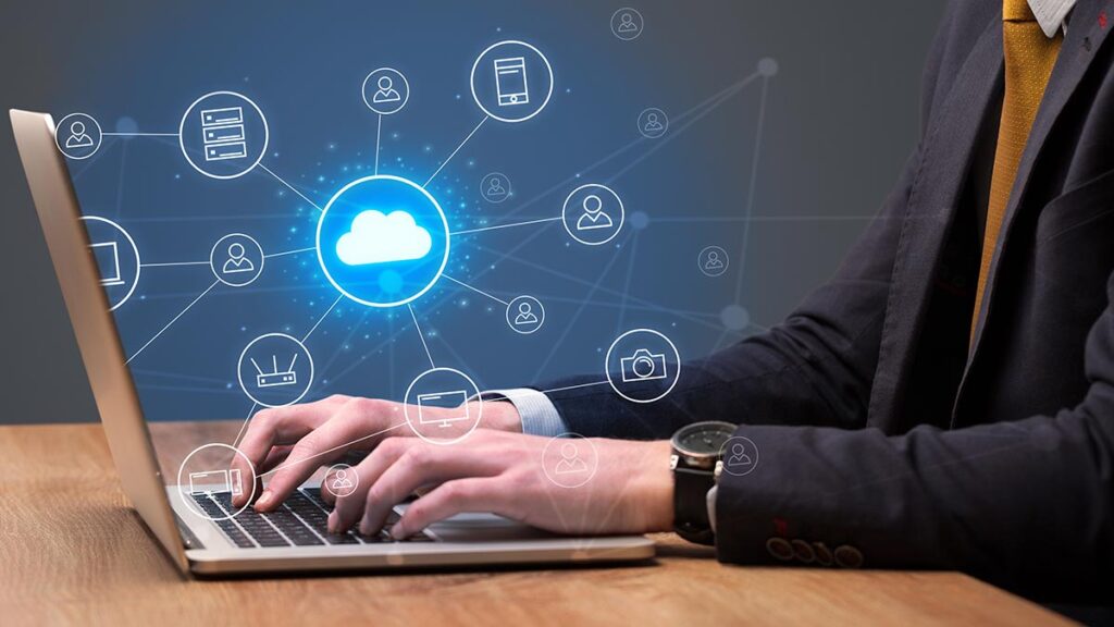 A person in a business suit typing on a laptop with a glowing digital cloud icon hovering above the keyboard. Lines radiate from the central cloud to various circular icons representing servers, mobile devices, users, cameras, and network connections, symbolising a cloud computing network.